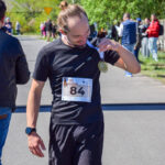 Mężczyzna w czarnej koszulce z numerem 84 na szyi trzyma medal. Tło to zielone drzewa i niebo. Grupa ludzi rozmawia obok.