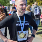 Młody mężczyzna z medalem stoi na ulicy. Ma czarną koszulkę i numer startowy. W tle są ludzie i zielone drzewa.