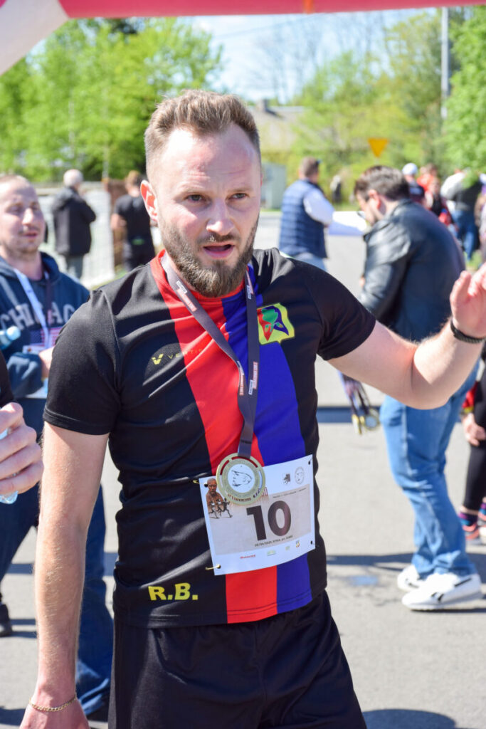 Mężczyzna w sportowej koszulce z medalem na tle uczestników wydarzenia. Ubrany w czarno-czerwono-niebieską koszulkę. W słoneczny dzień na świeżym powietrzu.