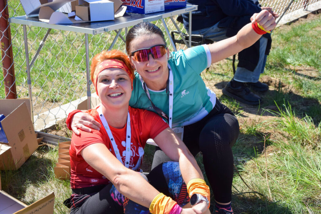 Dwie uśmiechnięte kobiety siedzą w trawie, ubrani w sportowe stroje. Jedna ma czerwoną koszulkę, druga niebieską. W tle metalowe ogrodzenie i pudełka.