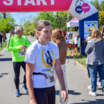 Na zdjęciu widać uczestników biegu pod łukiem z napisem START w tle. Kobieta w białej koszulce z medalem, widać również innych biegaczy i kibiców. Tło jest zielone, pełne drzew i ludzi.