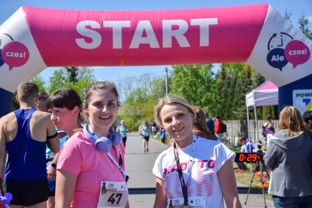 Grupa biegaczy przy starcie maratonu. Nad nimi różowy łuk z napisem START. Tło zielone drzewa i niebo.
