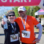 Na zdjęciu są dwie uśmiechnięte osoby w strojach sportowych z numerami startowymi. Kobieta trzyma butelkę z wodą, wokół której widać rozpryski, a mężczyzna ma na sobie pomarańczową koszulkę. W tle widać