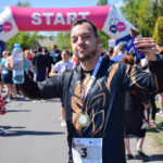 Nie wiem, kto jest na zdjęciu. Osoba z medalem, wodą i batonem stoi przed różowym banerem Start w otoczeniu biegaczy. Tło to zielone drzewa i niebieskie niebo.