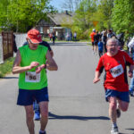 Na zdjęciu dwaj biegacze w jasnych koszulkach na wyścigu. Tło stanowi asfaltowa droga otoczona zielonymi drzewami. W tle widać kibiców oglądających bieg.