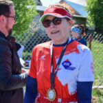 Nie wiem, kto jest na zdjęciu. Osoba w czerwonej koszulce z medalem rozmawia z innym uczestnikiem na tle zielonego ogrodu. Słoneczny dzień, metalowe ogrodzenie za nimi.