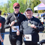 Dwóch mężczyzn w czarnych strojach z medalami na tle startowej bramy. Jasne, słoneczne niebo i zielone drzewa w tle. Uśmiechnięci, z numerami startowymi.