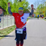 Mężczyzna w czerwono-białej koszulce finiszuje z uniesionymi rękami. Jasny dzień, zielone drzewa w tle. Numer startowy 89.