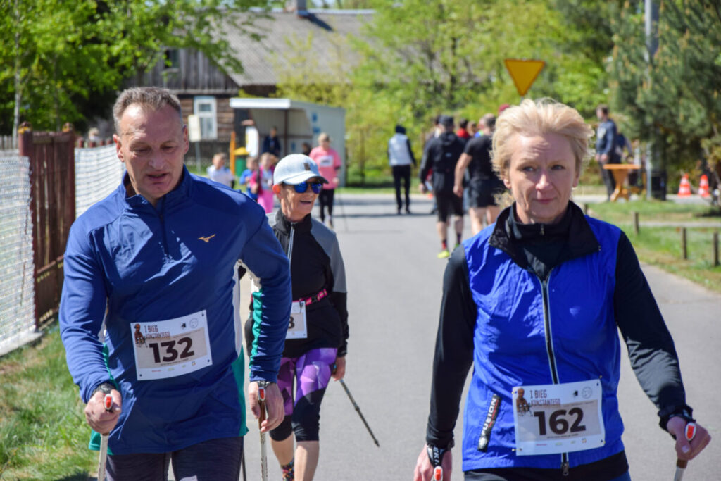 Uczestnicy marszu nordic walking z numerami startowymi idą ulicą. Domy i drzewa w tle, dominują niebieskie stroje. Jasne, słoneczne niebo.