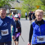 Uczestnicy marszu nordic walking z numerami startowymi idą ulicą. Domy i drzewa w tle, dominują niebieskie stroje. Jasne, słoneczne niebo.