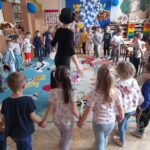 Dzieci w przedszkolu stoją w kółku na kolorowym dywanie. Nauczycielka w kapeluszu prowadzi zabawę. Tło zdobią barwne dekoracje.