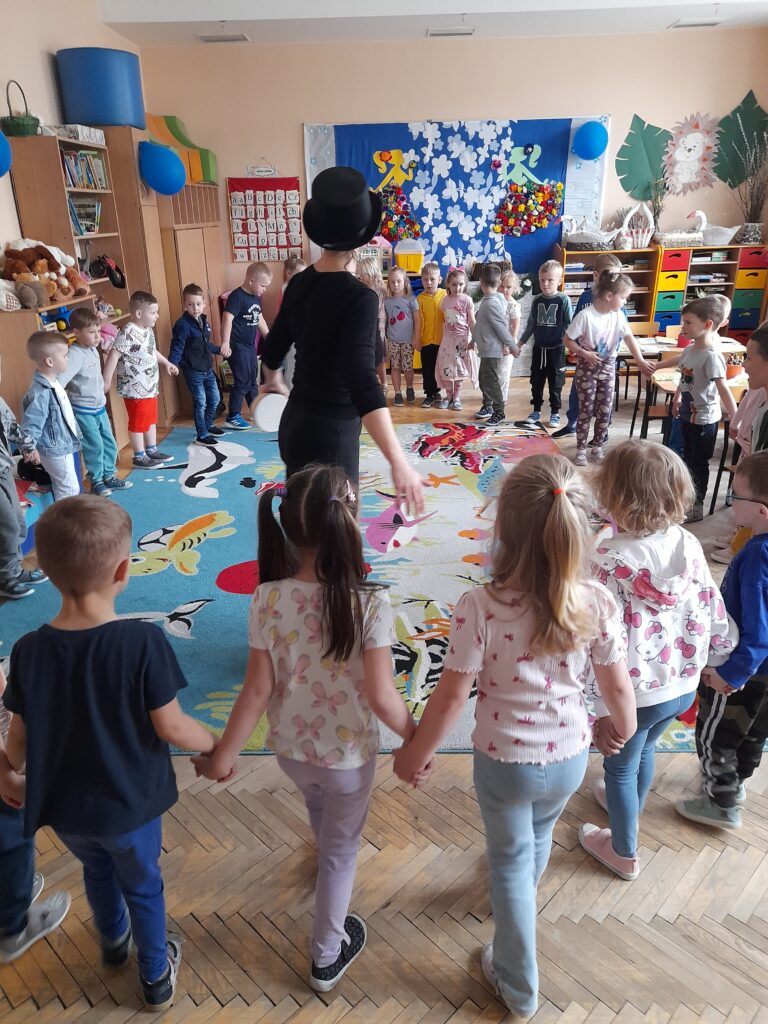 Dzieci w przedszkolu stoją w kółku na kolorowym dywanie. Nauczycielka w kapeluszu prowadzi zabawę. Tło zdobią barwne dekoracje.