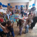 Dzieci bawią się w jasnej sali przedszkolnej z kolorowym dywanem i niebieskimi balonami. Na ścianie są dekoracje. Dzieci podnoszą ręce, uczestnicząc w zabawie.