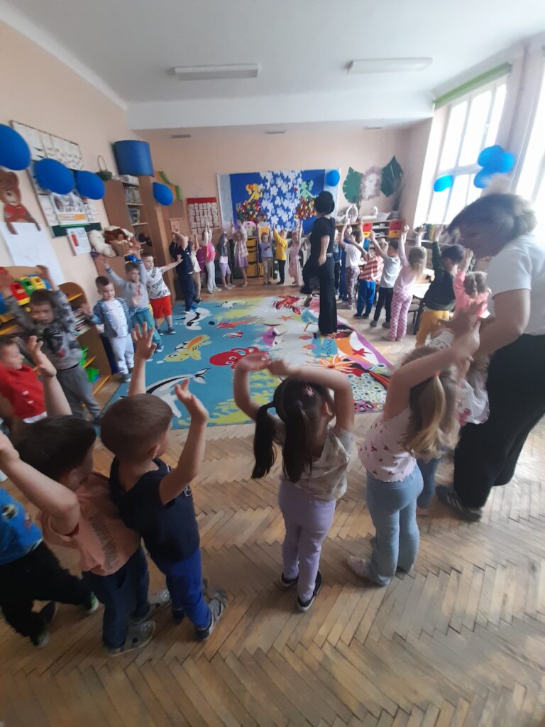 Dzieci bawią się w jasnej sali przedszkolnej z kolorowym dywanem i niebieskimi balonami. Na ścianie są dekoracje. Dzieci podnoszą ręce, uczestnicząc w zabawie.