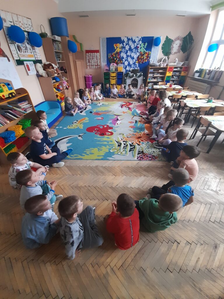 Grupa dzieci siedzi w kole na kolorowym dywanie w przedszkolu. W tle regały z zabawkami i książkami. Ściany ozdobione dekoracjami.