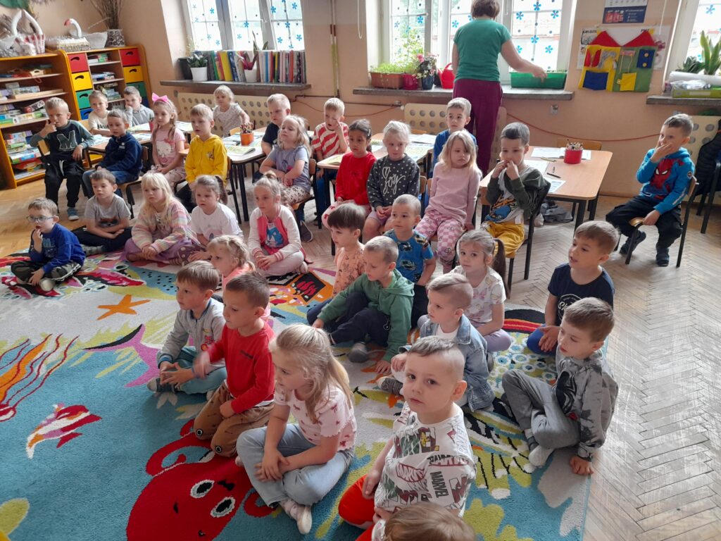 Grupa dzieci siedzi w przedszkolu na kolorowym dywanie. Za nimi niska pani przy oknie z niebieskimi dekoracjami. W tle widać regał z zabawkami i książkami.
