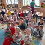 Grupa dzieci siedzi w przedszkolu na kolorowym dywanie. Za nimi niska pani przy oknie z niebieskimi dekoracjami. W tle widać regał z zabawkami i książkami.