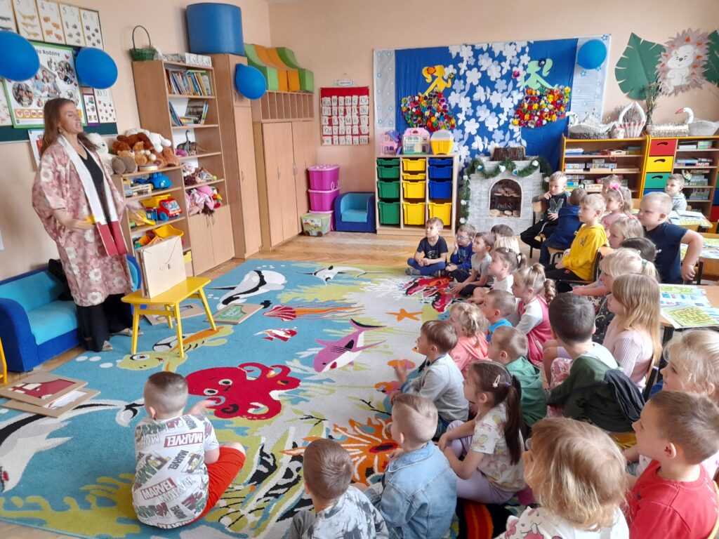Sala przedszkolna z kolorowym dywanem. Dzieci siedzą przed nauczycielką w kwiecistym stroju. Kolorowe ozdoby i zabawki na półkach.