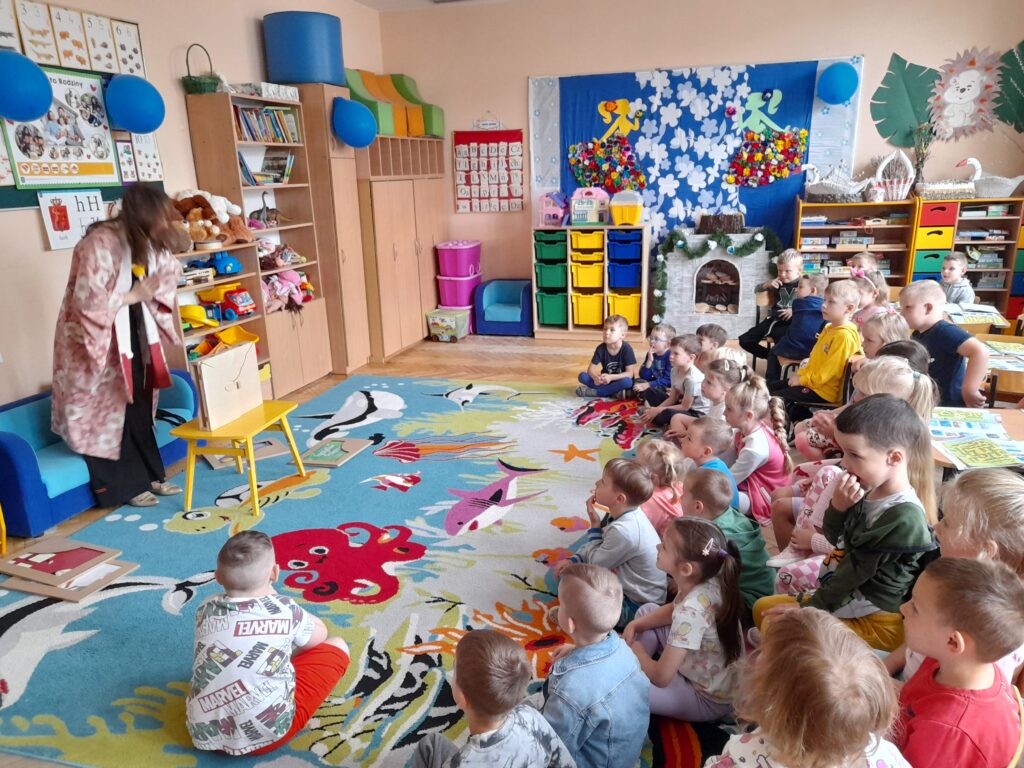 Dzieci siedzą na kolorowym dywanie w klasie, patrząc na nauczyciela. Jasne ściany z dekoracjami, półki pełne zabawek. Niebieskie i żółte meble podkreślają radosny wystrój.