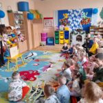 Dzieci siedzą na kolorowym dywanie w klasie, patrząc na nauczyciela. Jasne ściany z dekoracjami, półki pełne zabawek. Niebieskie i żółte meble podkreślają radosny wystrój.