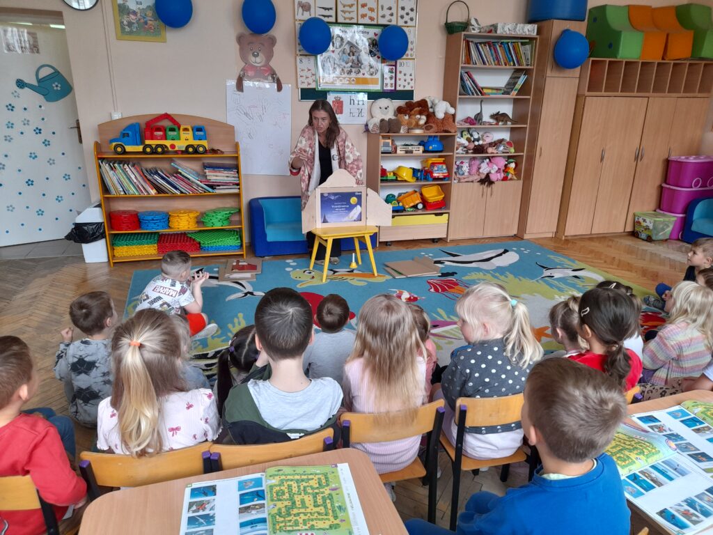 Na zdjęciu widać dzieci siedzące na kolorowym dywanie w przedszkolu. Przed nimi nauczycielka czyta książkę, za nią półki z zabawkami. Ściany są ozdobione plakatami i rysunkami.