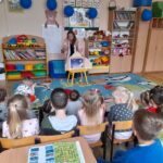 Na zdjęciu widać dzieci siedzące na kolorowym dywanie w przedszkolu. Przed nimi nauczycielka czyta książkę, za nią półki z zabawkami. Ściany są ozdobione plakatami i rysunkami.