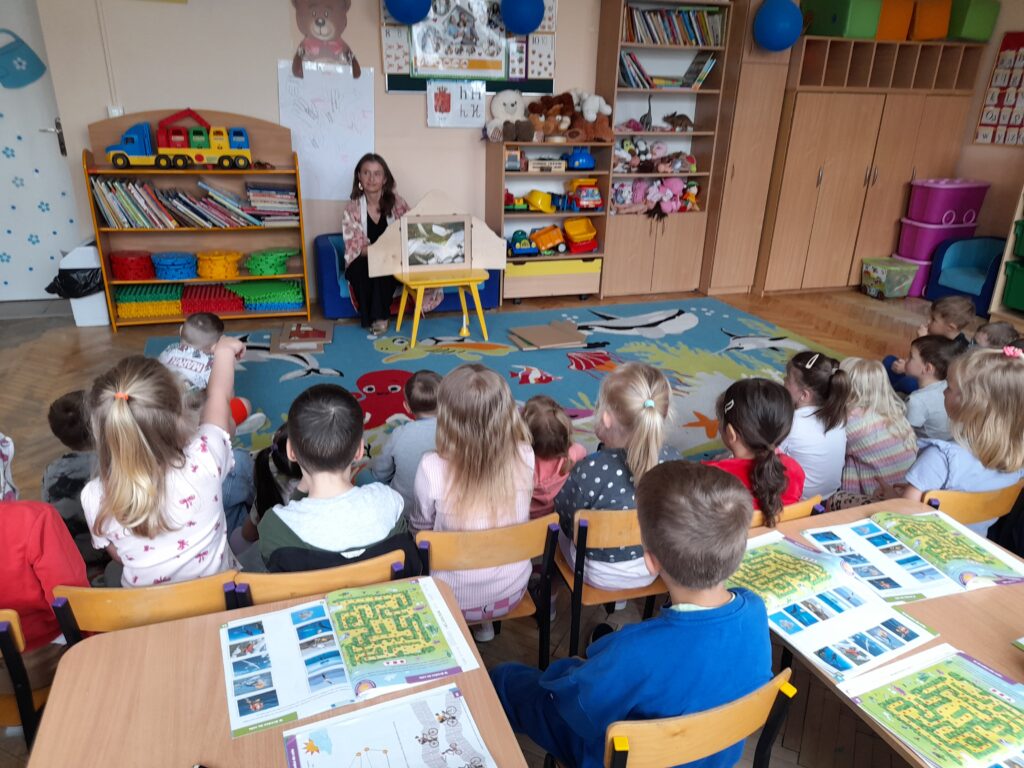 Dzieci siedzą na kolorowym dywanie w sali przedszkolnej. Nauczycielka pokazuje coś na małym stoliku. W tle regały z książkami i zabawkami.