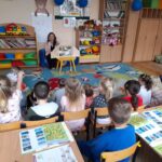Dzieci siedzą na kolorowym dywanie w sali przedszkolnej. Nauczycielka pokazuje coś na małym stoliku. W tle regały z książkami i zabawkami.