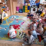 Grupa dzieci siedzi na kolorowym dywanie z motywami ryb. Kobieta pokazuje im książkę przy drewnianej tablicy. W tle kolorowe szafki i zabawki.