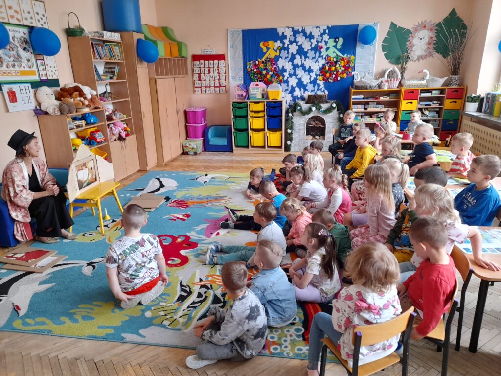 Na zdjęciu grupa dzieci w przedszkolu słucha opowieści. Kolorowy dywan z motywem zwierząt i regały z zabawkami w tle. Nauczycielka po lewej w kapeluszu przy tablicy.