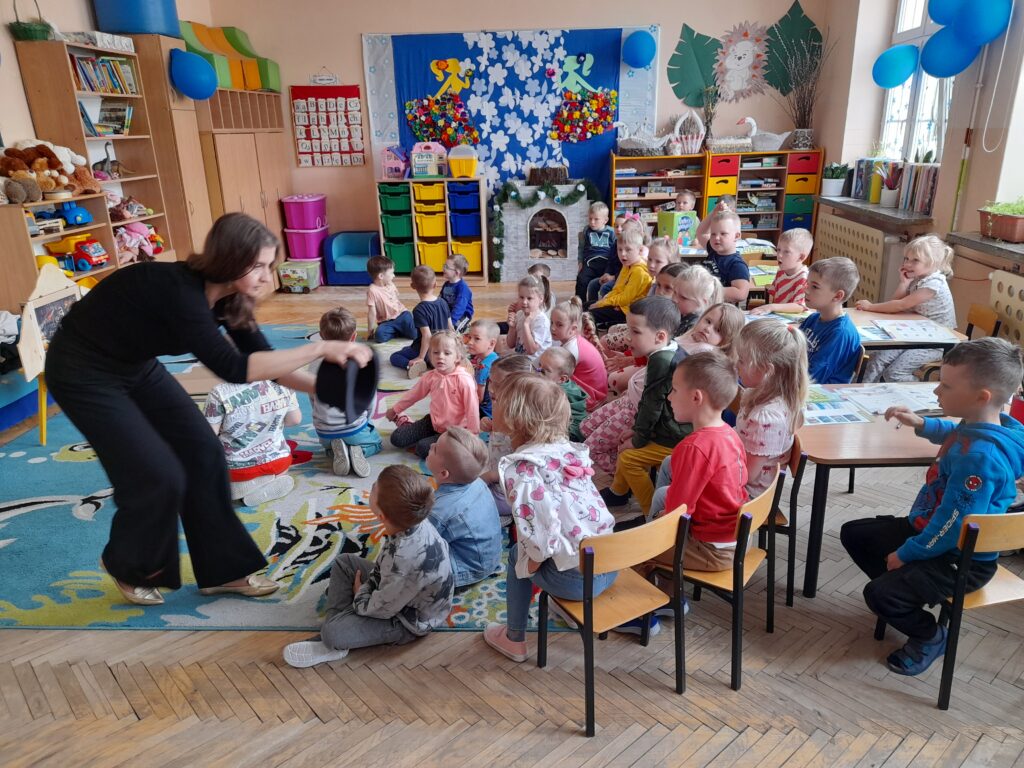 Grupa dzieci siedzi na kolorowym dywanie, uważnie patrząc na kobietę. W tle regały z zabawkami i kolorowe pudełka. Sala ozdobiona jest niebieskimi balonami i rysunkami.