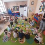 Dzieci siedzą w kręgu na kolorowym, zielonym dywanie z rysunkami zwierząt w jasnej sali przedszkolnej. Na ścianach wiszą tablice edukacyjne. Pomieszczenie jest udekorowane dziecięcymi zabawkami i plakatami.