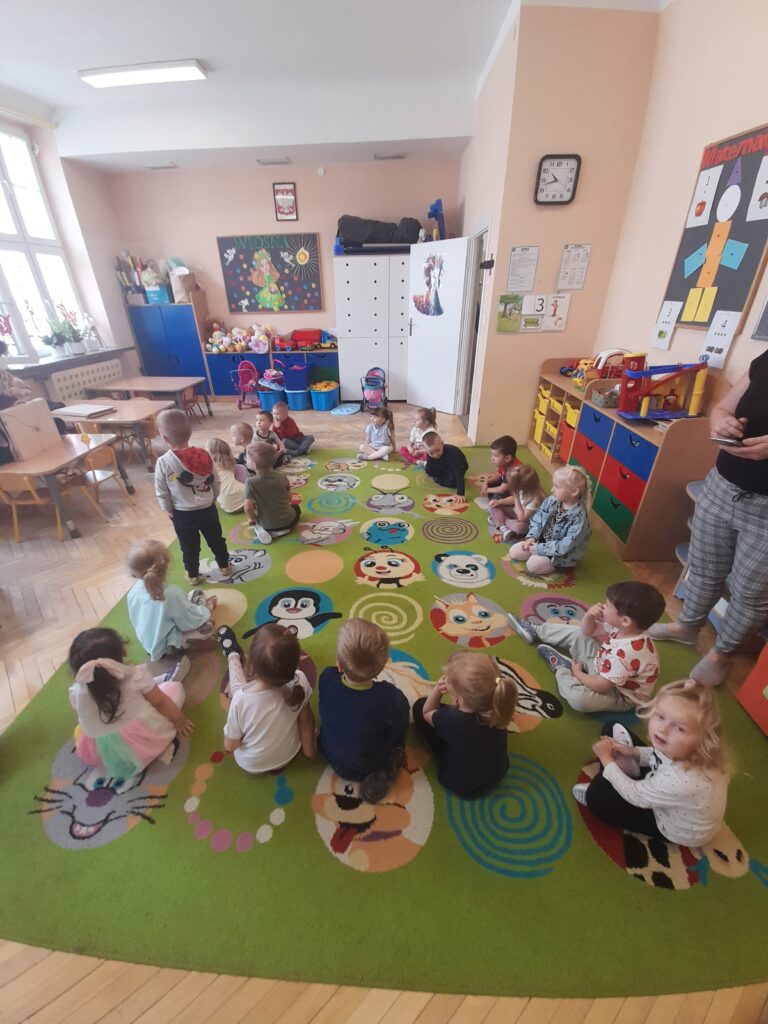 Dzieci siedzą w kręgu na kolorowym, zielonym dywanie z rysunkami zwierząt w jasnej sali przedszkolnej. Na ścianach wiszą tablice edukacyjne. Pomieszczenie jest udekorowane dziecięcymi zabawkami i plakatami.