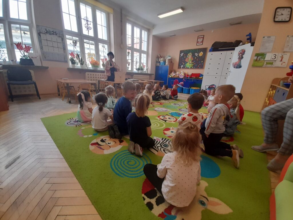 Grupa dzieci siedzi na kolorowym dywanie w przedszkolnej sali. Jasne ściany i duże okna wpuszczają dużo światła. Nauczycielka stoi z przodu pokoju, dekoracje w tle.
