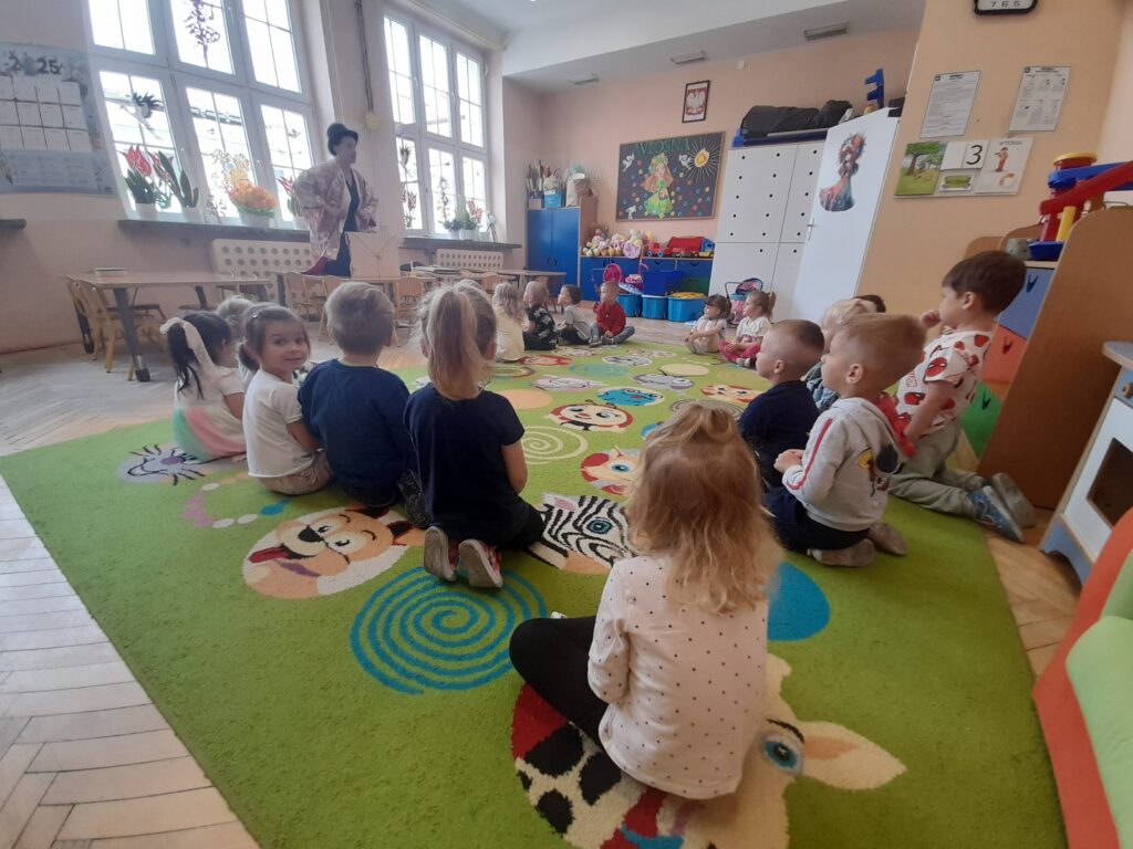 Dzieci siedzą na kolorowym dywanie w przedszkolu. Tło stanowi jasna ściana z oknami. Nauczycielka stoi z przodu.