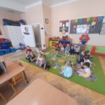 Dzieci siedzą na kolorowym dywanie w przedszkolu. Ściany udekorowane są plakatami i rysunkami. Tło jasne, meble drewniane.