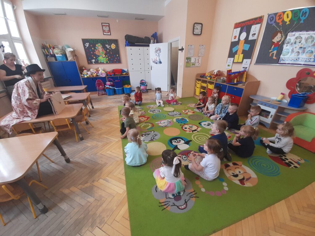 W przedszkolu dzieci siedzą na dywanie z kolorowymi wzorami. Nauczyciel w kapeluszu pokazuje książkę. W tle jasne ściany i regały z zabawkami.