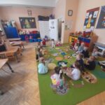 W przedszkolu dzieci siedzą na dywanie z kolorowymi wzorami. Nauczyciel w kapeluszu pokazuje książkę. W tle jasne ściany i regały z zabawkami.