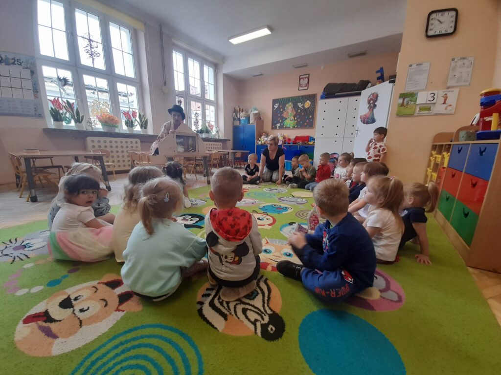 Sala przedszkolna z dziećmi siedzącymi na kolorowym dywanie. Na tle jasnych ścian i dużych okien są szafki i zabawki. Nauczyciel prezentuje coś na stoliku.