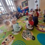Dzieci siedzą w kręgu na kolorowym dywanie w sali przedszkolnej. W tle jasne okna i dekoracje na ścianach. Dywan ma wzory zwierząt.