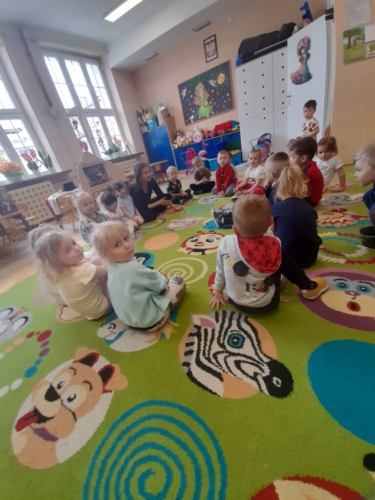Dzieci siedzą w kręgu na kolorowym dywanie w sali przedszkolnej. W tle jasne okna i dekoracje na ścianach. Dywan ma wzory zwierząt.