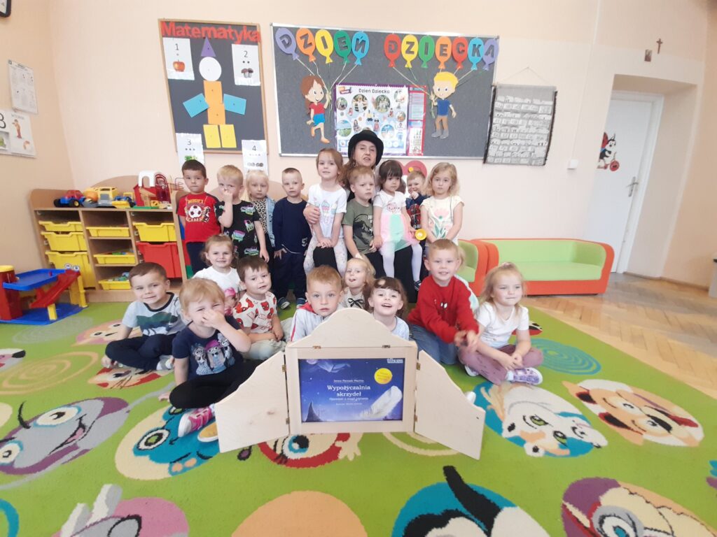 Grupa uśmiechniętych dzieci siedzi na kolorowym dywanie z postaciami. W tle szafki i ozdoby ścienne. Dzieci pozują przy drewnianej książce.