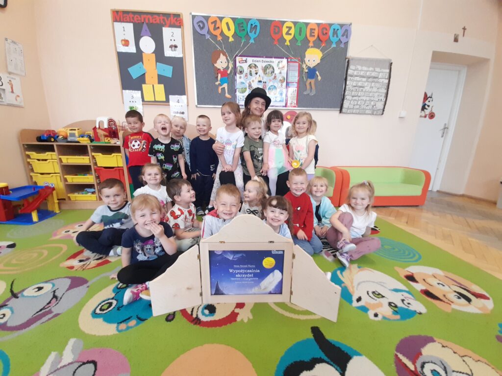 Grupa dzieci na kolorowym dywanie w przedszkolu, w tle jasna ściana z dekoracjami i półki z zabawkami.