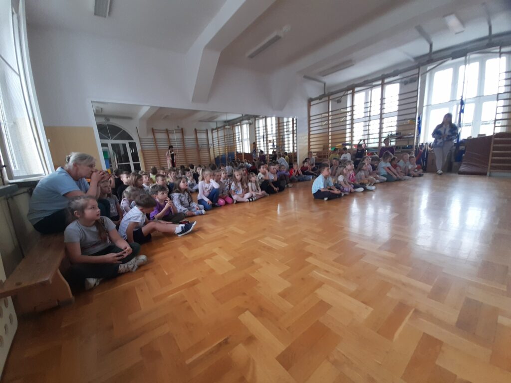 Dzieci siedzą na drewnianej podłodze w jasnej sali gimnastycznej. Na lewo starsza kobieta z aparat. Ściany mają drabinki gimnastyczne.