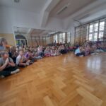 Dzieci siedzą na podłodze w sali gimnastycznej, z drewnianą podłogą i drabinkami na ścianie. Kolory są stonowane, z przewagą jasnych odcieni. Światło wpada przez duże okna, tworząc jas