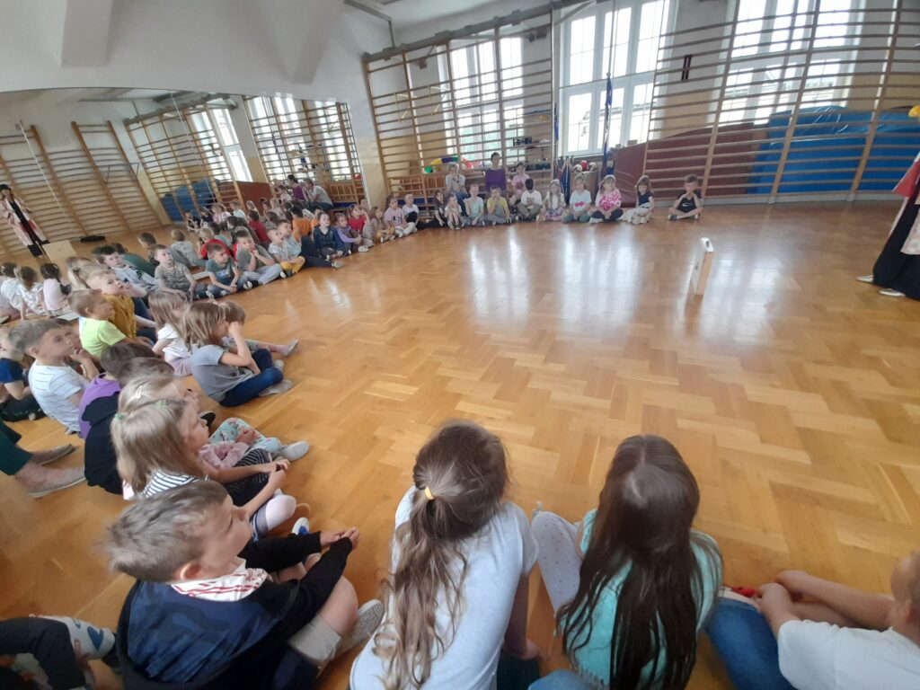 Grupa dzieci siedzi na jasnej podłodze sali gimnastycznej. W tle lustra i drabinki, światło wpada przez duże okna. Dzieci są skupione, patrzą na przedmiot pośrodku.