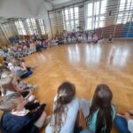 Grupa dzieci siedzi na jasnej podłodze sali gimnastycznej. W tle lustra i drabinki, światło wpada przez duże okna. Dzieci są skupione, patrzą na przedmiot pośrodku.