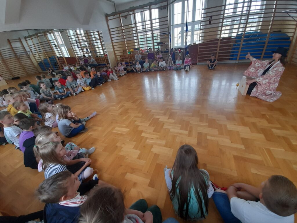 Dzieci siedzą w dużym jasnym pomieszczeniu na podłodze z drewna. Na środku starszy mężczyzna w różowej szacie wykonuje przedstawienie. W tle duże okna i drabinki gimnastyczne.