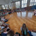 Dzieci siedzą w dużym jasnym pomieszczeniu na podłodze z drewna. Na środku starszy mężczyzna w różowej szacie wykonuje przedstawienie. W tle duże okna i drabinki gimnastyczne.