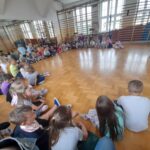 Dzieci siedzące w kręgu na drewnianej podłodze sali gimnastycznej. Sala ma jasne ściany i drabinki przy ścianach. Z prawej strony osoba pokazuje obrazki.